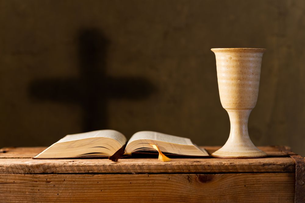 Becher und Bibel auf Tisch vor dem Schatten eines Kreuzes an der Wand